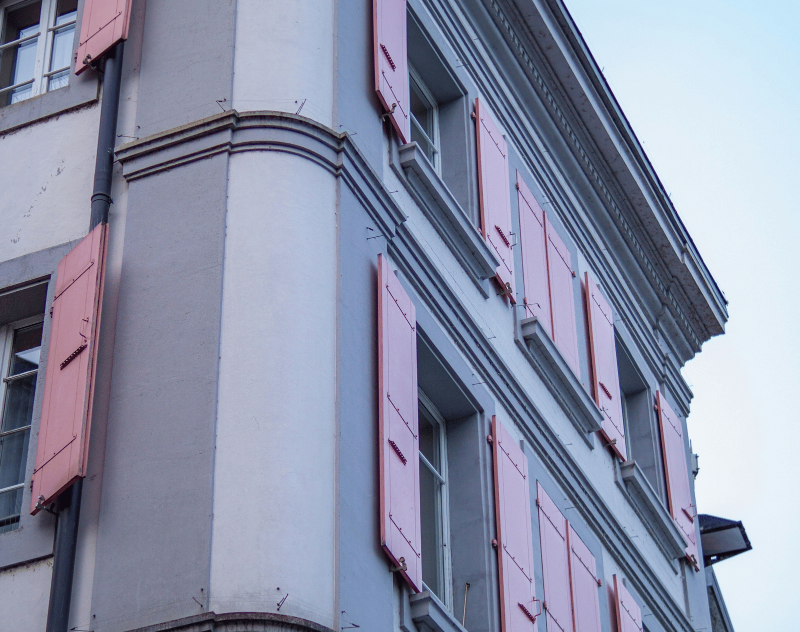 A low-angle view of a grey stone building featuring traditional pink window shutters.