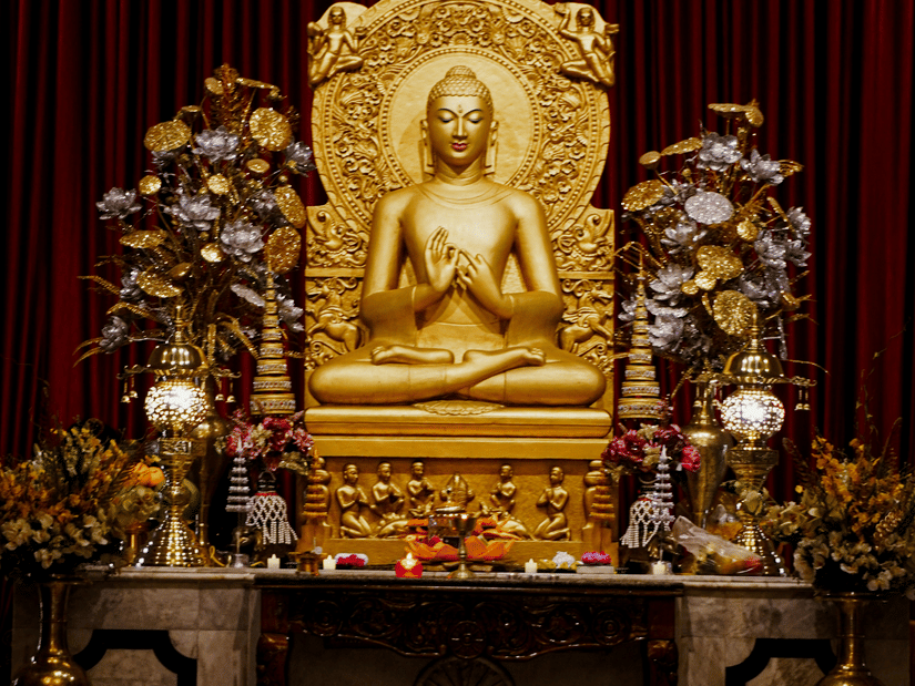 A gold-coloured statue of Buddha seated in a shrine against a dark red curtain | HHI Varanasi