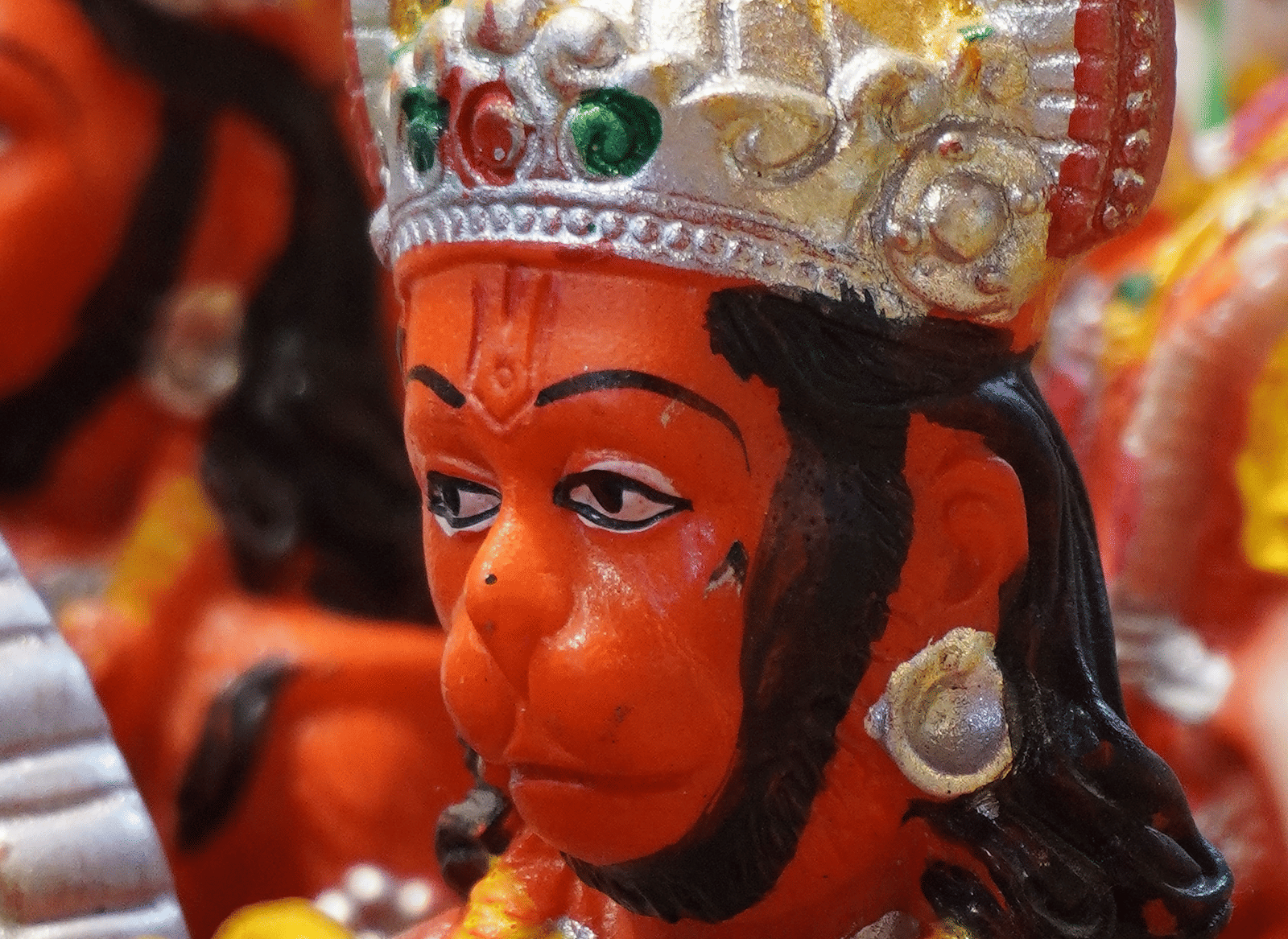 Close-up of a painted orange Lord Hanuman idol wearing a gold and silver crown with green and red jewels, with more idols blurred in the background.