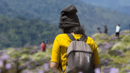 Traveller hiking through blooming wildflowers on a scenic mountain trail during the monsoon, surrounded by lush green hills and panoramic valley views.