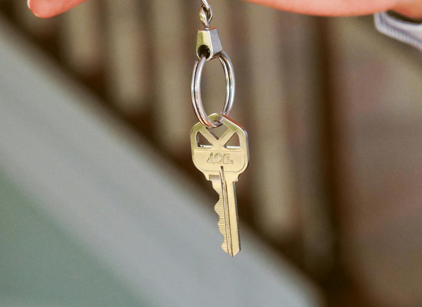 A hand holds up a single silver house key, dangling from the thumb and forefinger, against a blurred background of wooden stairs.