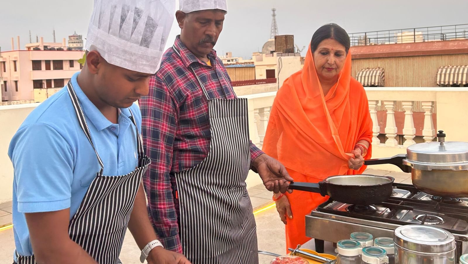 A woman wearing a saree and two men wearing chef hats showcasing cooking on the rooftop - Dileep Kothi, Jaipur