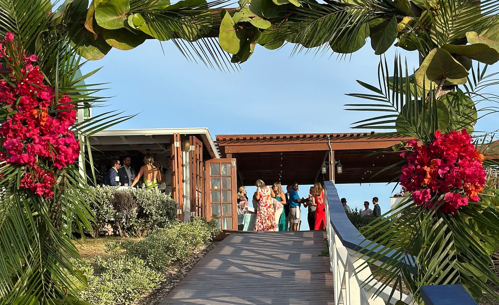 An wooden pathway leading to the cottage style courtyard where people have gathered to celebrate a wedding. The entrance of the decorated with branches, leaves, and flowers. 