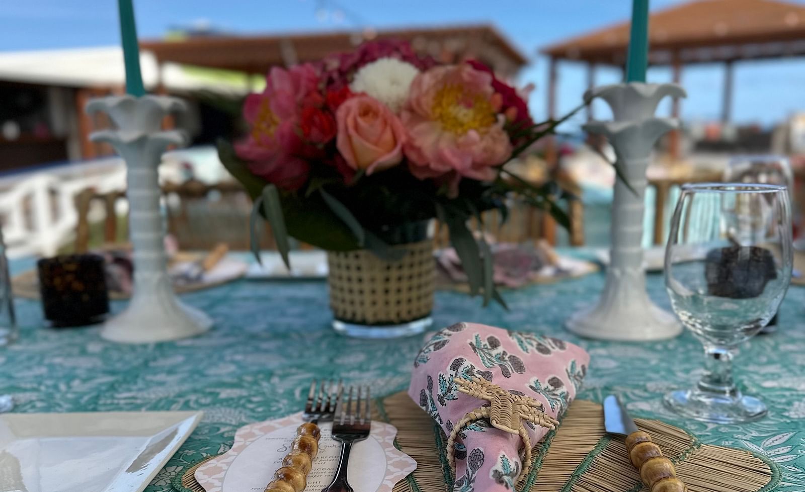 A dining table at the beachside featuring a bouquet the centre of the table, a table mat where knife and forks are placed, along with a wine glass and candle stands. 