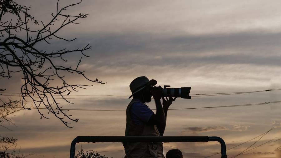 A man clicking pictures using a camera, standing on a jeep, with a clear sky in the background -Daksh Eden Greenz, Sasan Gir