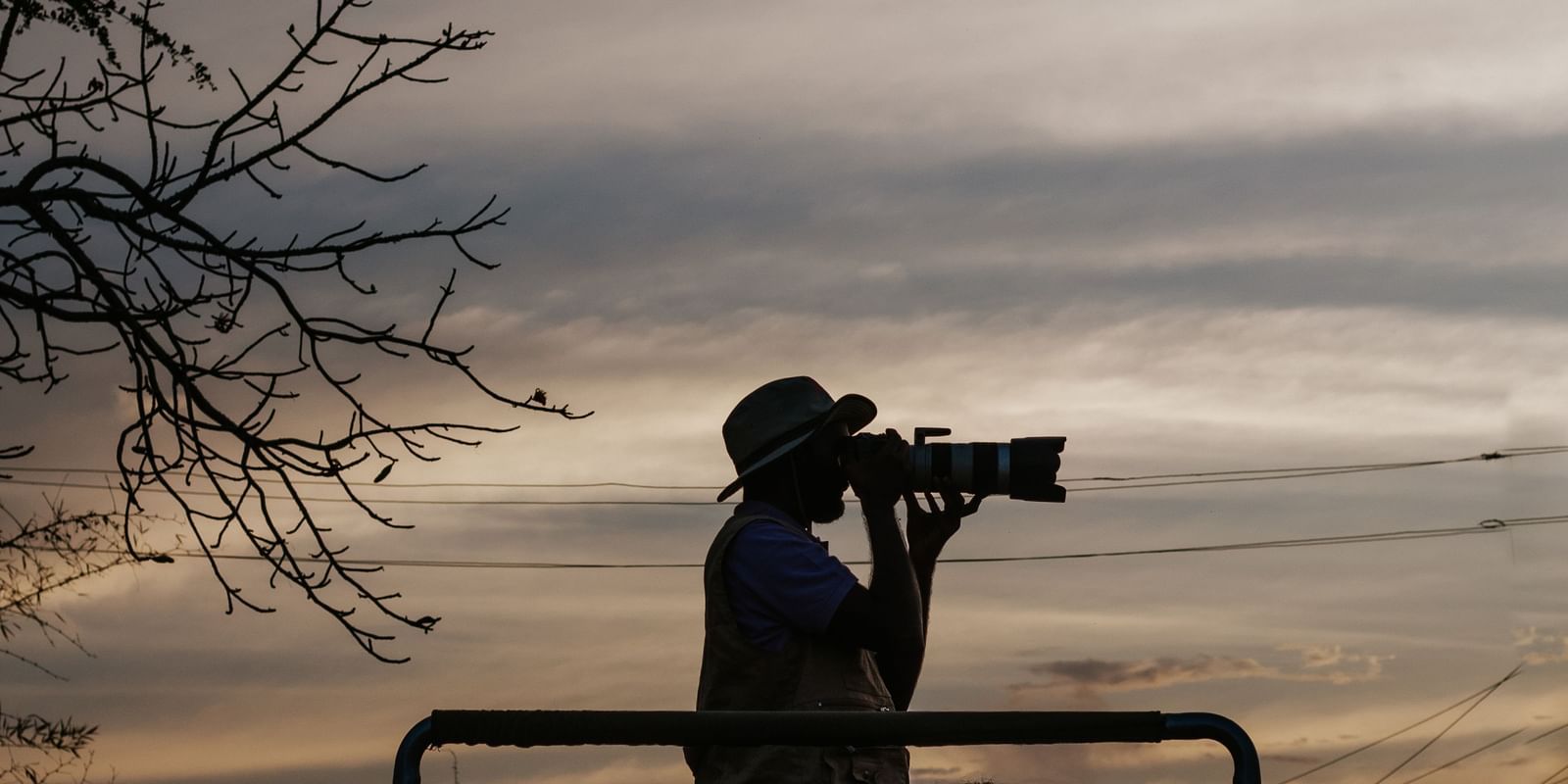 A man clicking pictures using a camera, standing on a jeep, with a clear sky in the background -Daksh Eden Greenz, Sasan Gir