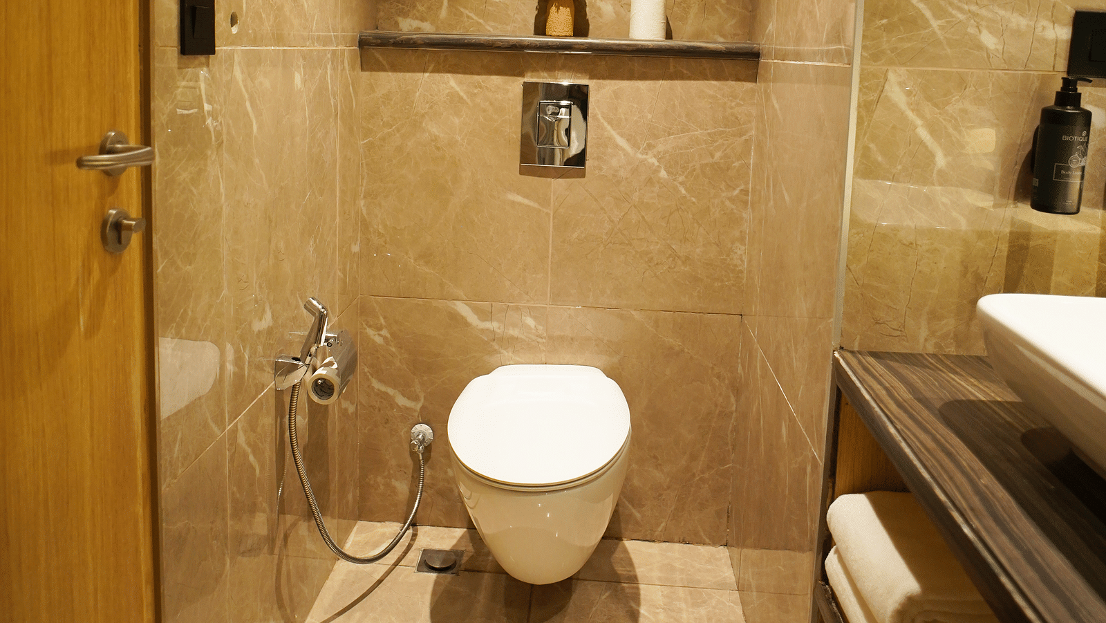 Compact bathroom featuring toilet, bidet spray, and wooden door - Ramada Encore by Wyndham Gurugram Dwarka Expressway