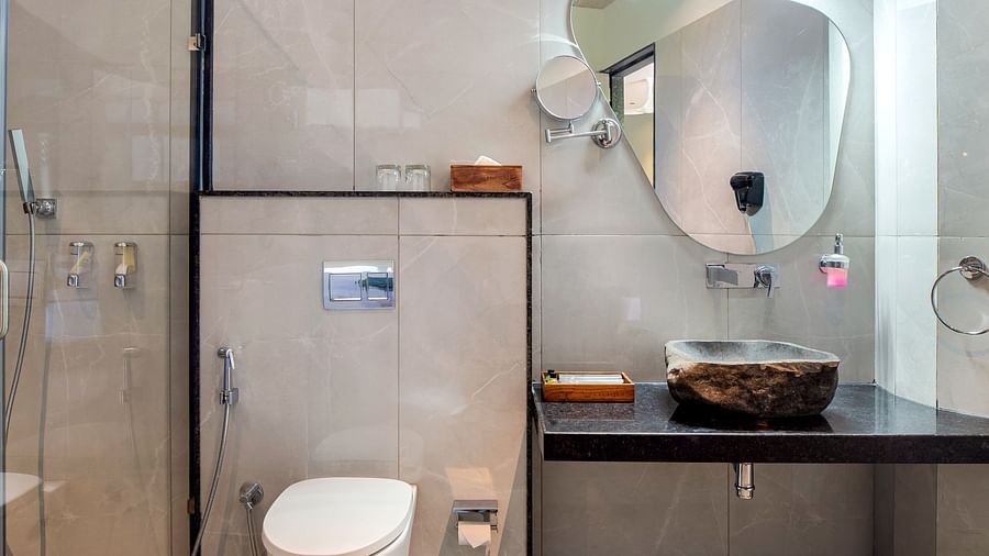 A bathroom of Club Suite with Valley View Sitout showcasing a toilet, a sink with a mirror, and a shower area enclosed by a glass door at The Qualia Resort Club & Brewery, Udaipur.