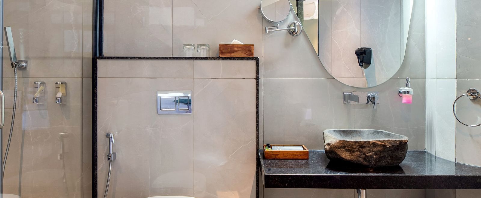 A bathroom of Club Suite with Valley View Sitout showcasing a toilet, a sink with a mirror, and a shower area enclosed by a glass door at The Qualia Resort Club & Brewery, Udaipur.