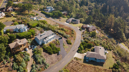 An aerial view of scattered homes built among rolling, tree-covered hills and coastal scrubland.