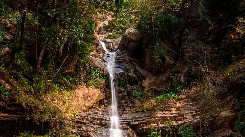 A picturesque waterfall gracefully flows over layered rocks into a clear, calm pool of water, surrounded by lush green vegetation and natural rock formations, creating a serene natural oasis