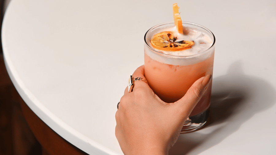 Two people sitting at a round white table, each holding a different cocktail—one in a short glass garnished with citrus peel, and the other in a tall stemmed glass with a frothy top and a lemon twist - Ananta Spa and Resort, Pushkar.