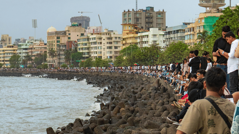 A vertical shot of Marine Drive, Mumbai, with people sitting on the promenade wall beside a shoreline of dark tetrapods and a city skyline under a grey sky.