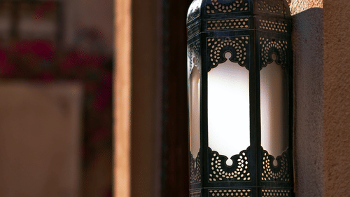 A close-up shot of an elegant hanging lantern with intricate patterns, set against a carved, reddish-brown wall.
