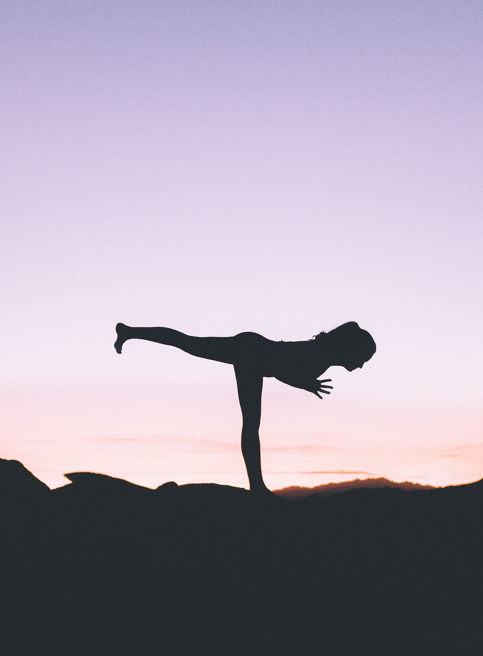 a silhouette of a woman doing a yoga pose amidst nature with pink and purple hued sky in the background