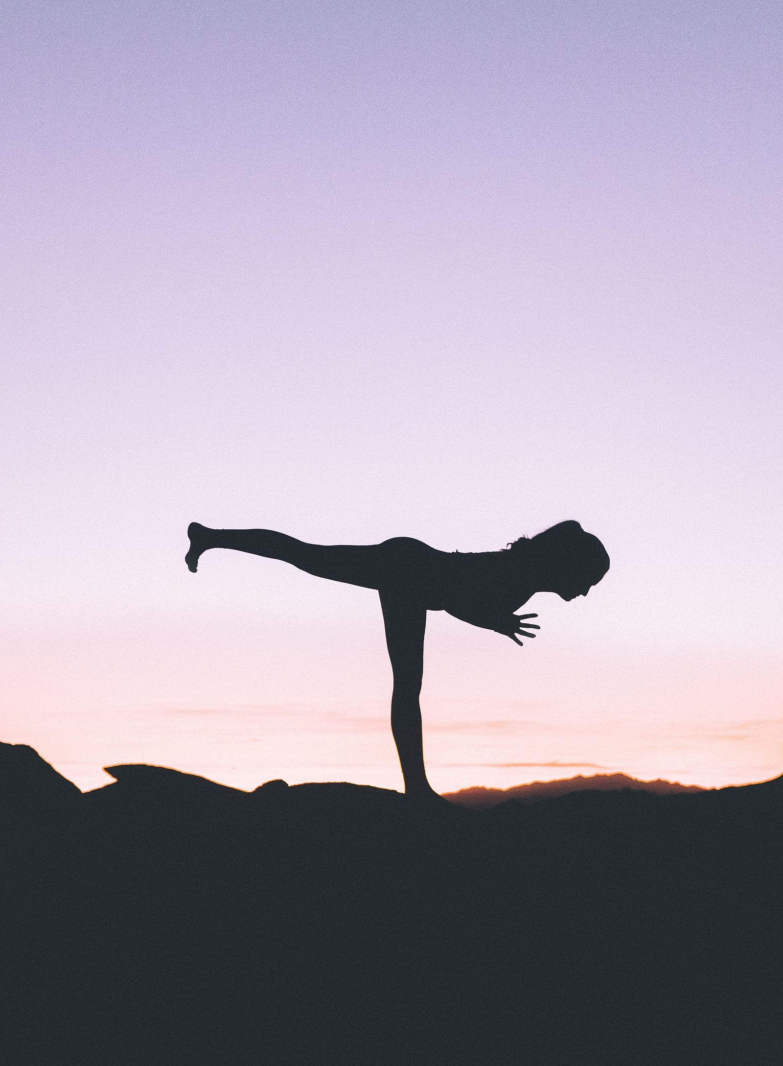 a silhouette of a woman doing a yoga pose amidst nature with pink and purple hued sky in the background