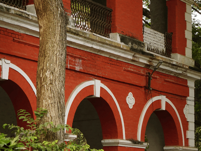 A vibrant red brick building with grand white arches partially framed by lush green foliage