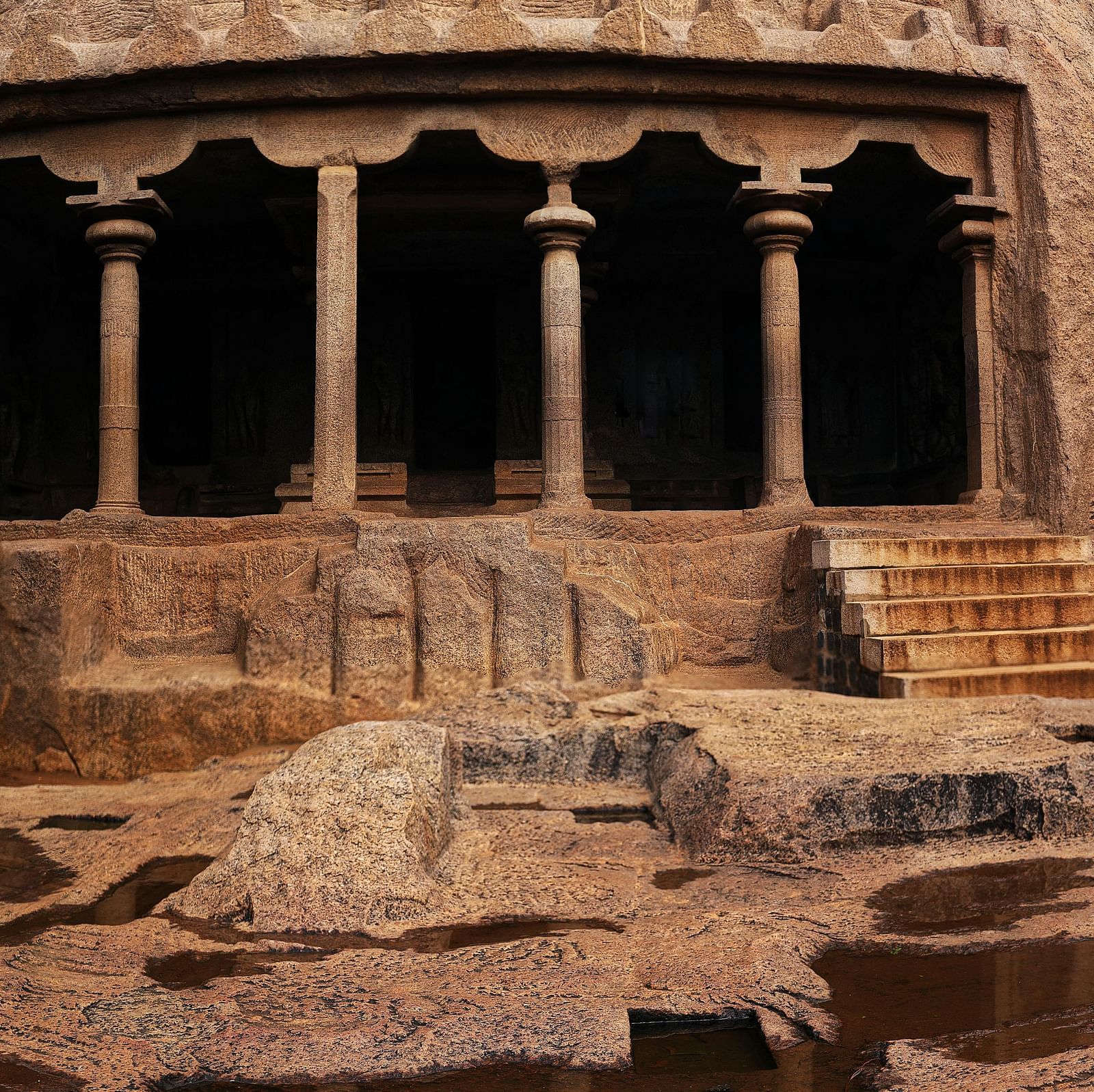 Ancient rock-cut featuring stone pillars, carved walls, and natural rock formations with shallow water pools in the foreground.