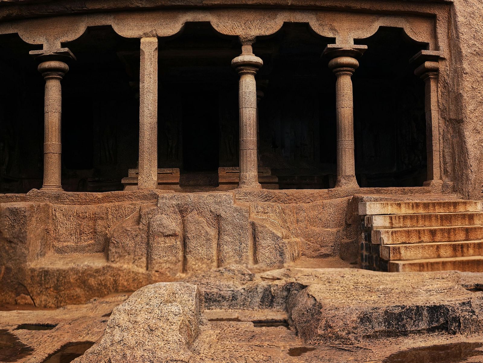 Ancient rock-cut featuring stone pillars, carved walls, and natural rock formations with shallow water pools in the foreground.