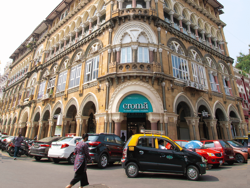View of a classic Mumbai building with parked cars and street life under a clear sky.