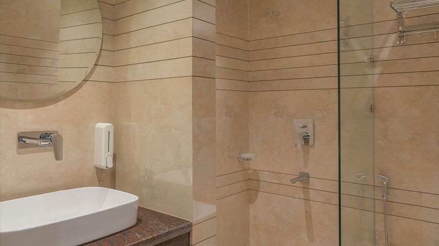 A bathroom with a wash basin above a round wall-mounted mirror and a glass separating the shower area | Hotel Ganga Maiya