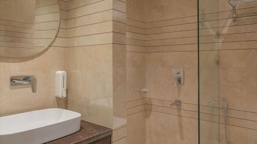A bathroom with a wash basin above a round wall-mounted mirror and a glass separating the shower area | Hotel Ganga Maiya