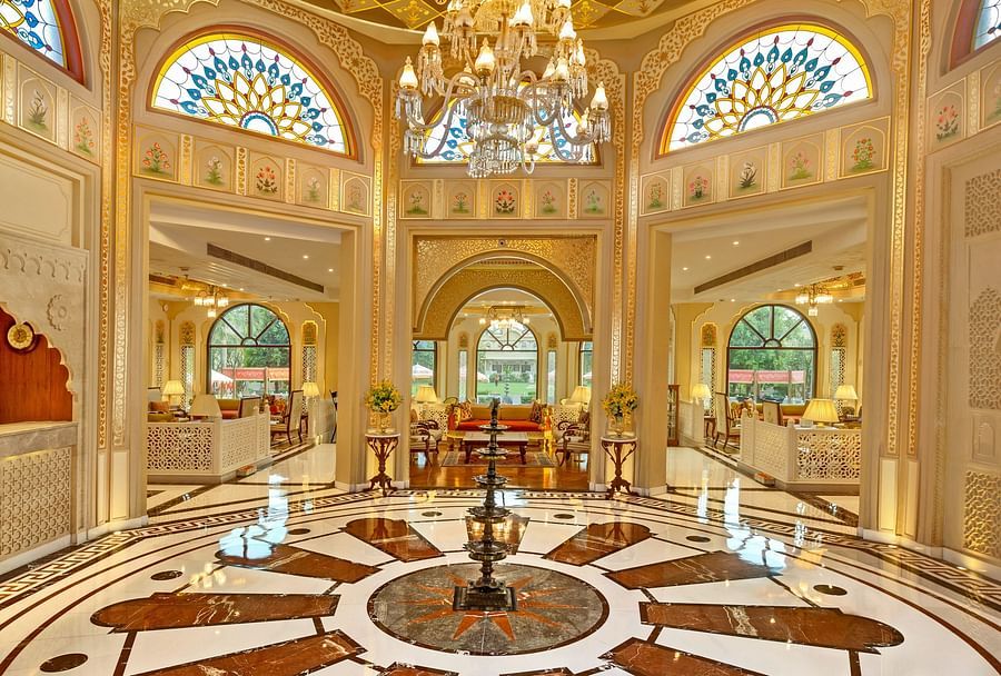Interior of a luxurious room with ornate gold and cream patterned ceiling, large arched windows, and a decorative brown and white floor design, illuminated by a grand chandelier.