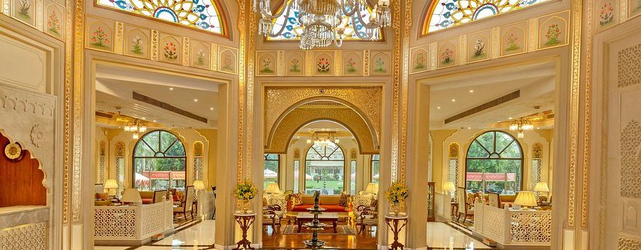 Interior of a luxurious room with ornate gold and cream patterned ceiling, large arched windows, and a decorative brown and white floor design, illuminated by a grand chandelier.