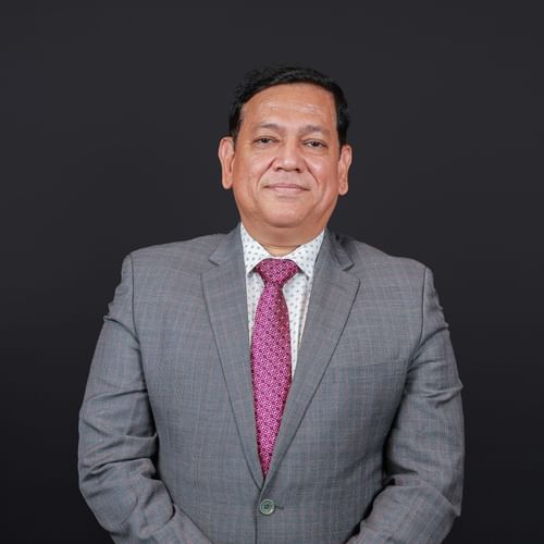Professional portrait of a man in a grey suit with a pink tie, standing confidently against a black background.