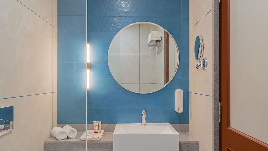 A spacious bathroom with a round wall-mounted mirror above a wash basin and a glass partition separating the shower area | Hotel Ganga Maiya