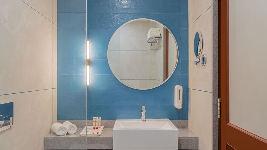 A spacious bathroom with a round wall-mounted mirror above a wash basin and a glass partition separating the shower area | Hotel Ganga Maiya