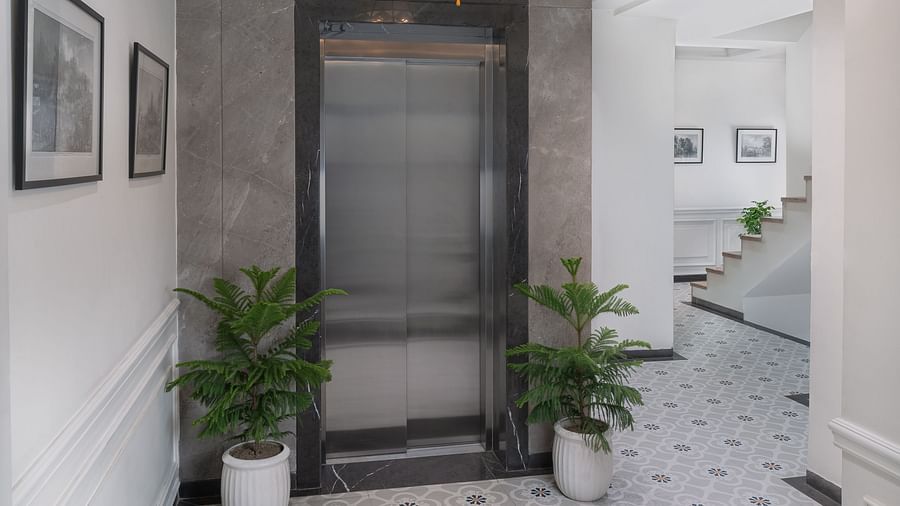 A corridor leads to the lift entrance, with two plants on each side, wall-mounted photo frames, and a chandelier overhead | Hotel Ganga Maiya