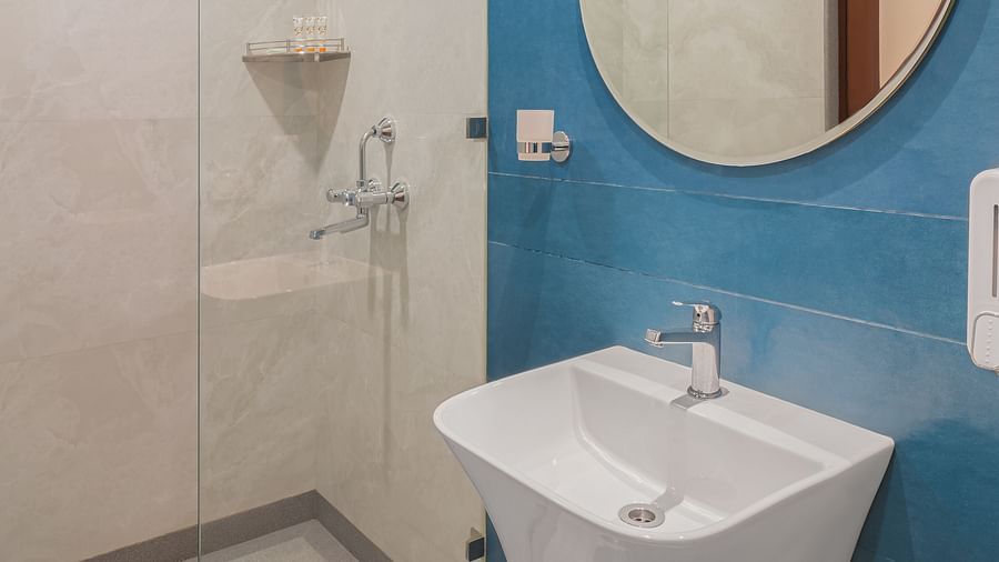 A bathroom with a wash basin above a round wall-mounted mirror and a glass separating the shower area | Hotel Ganga Maiya