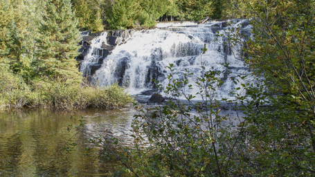 A multi-tiered waterfall cascading down rock steps into a calm stream surrounded by dense green forest trees.