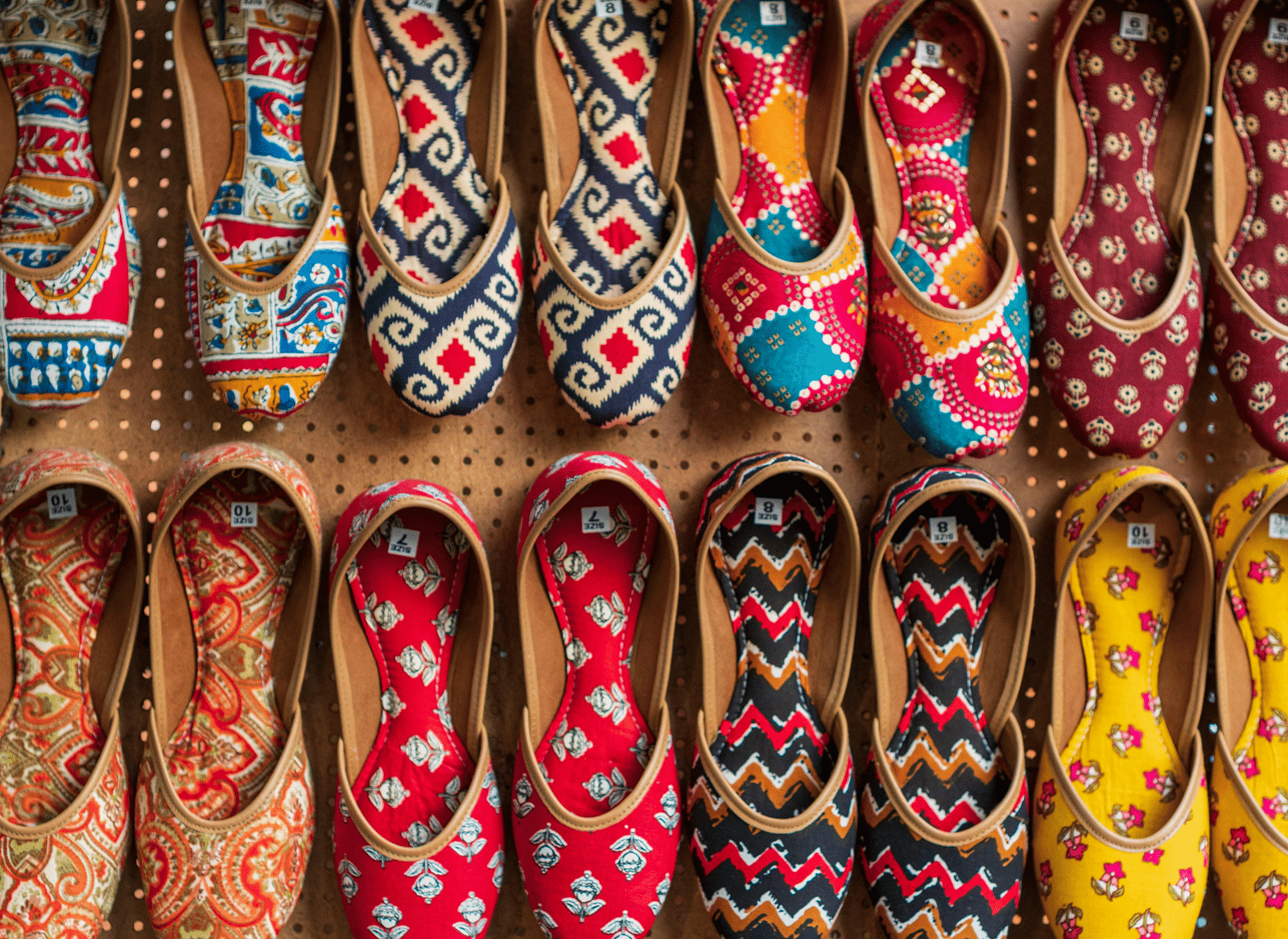 A vibrant display showcases rows of traditional Indian shoes, or juttis, featuring colourful embroidery and beadwork, arranged neatly with varying patterns and hues against a neutral backdrop.