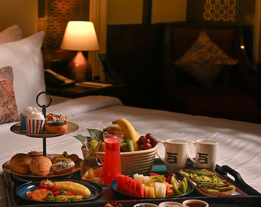 Luxurious breakfast spread served on a bed in a warmly lit hotel room, featuring a variety of dishes including fresh fruit, pastries, juice, and coffee - Ananta Spa and Resort, Pushkar.