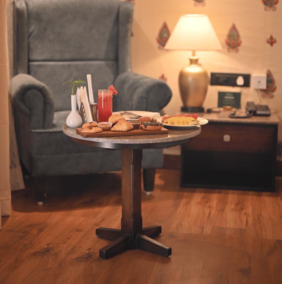 A Premium Tent featuring a sofa chair, a centre table which has a tray with croissant, cookies, doughnuts, a glass of juice and near to it there is a bedside table  with lamp and a telephone - The Baagh Ananta Elite, Ranthambore
