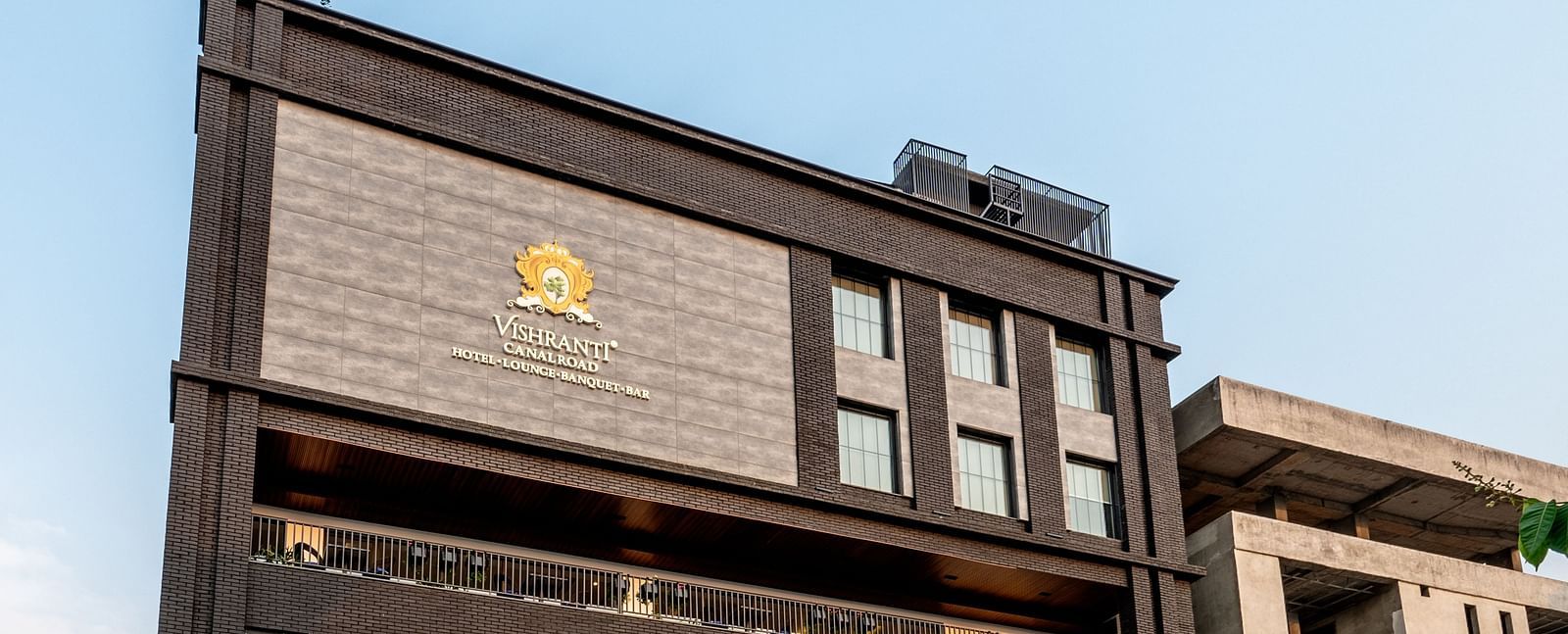 Side shot of the facade of Vishranti Hotel Dehradun showcasing its logo engraved on the wall taken during the day.