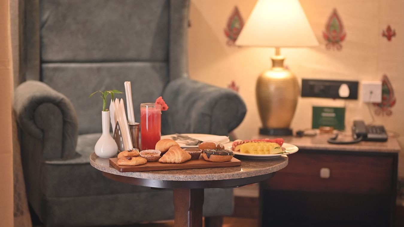A Premium Tent featuring a sofa chair, a centre table which has a tray with croissant, cookies, doughnuts, a glass of juice and near to it there is a bedside table  with lamp and a telephone - The Baagh Ananta Elite, Ranthambore