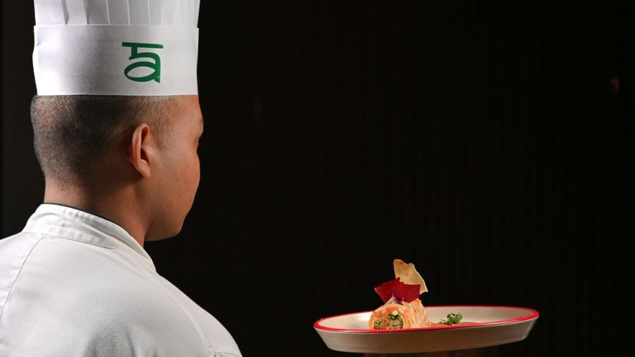 A chef wearing a tall white hat and white uniform is seen from behind, holding a plated gourmet dish with careful presentation, against a dark background - Ananta Spa and Resort, Pushkar.