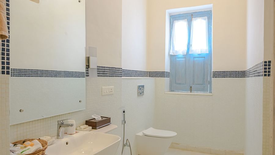 A bathroom with white walls and fixtures, a sink, toilet and a small window with a blind in the background. - Tijara Fort-Palace - 19th Century, Alwar