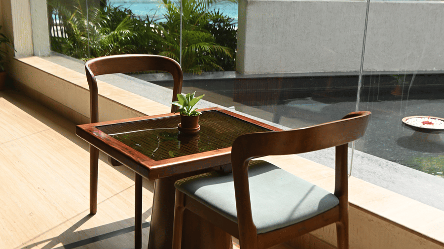 A wooden table and chair placed near large glass windows at Vedic Village Spa Resort overlooking a sunlit garden and pool area with lush green trees visible outside.
