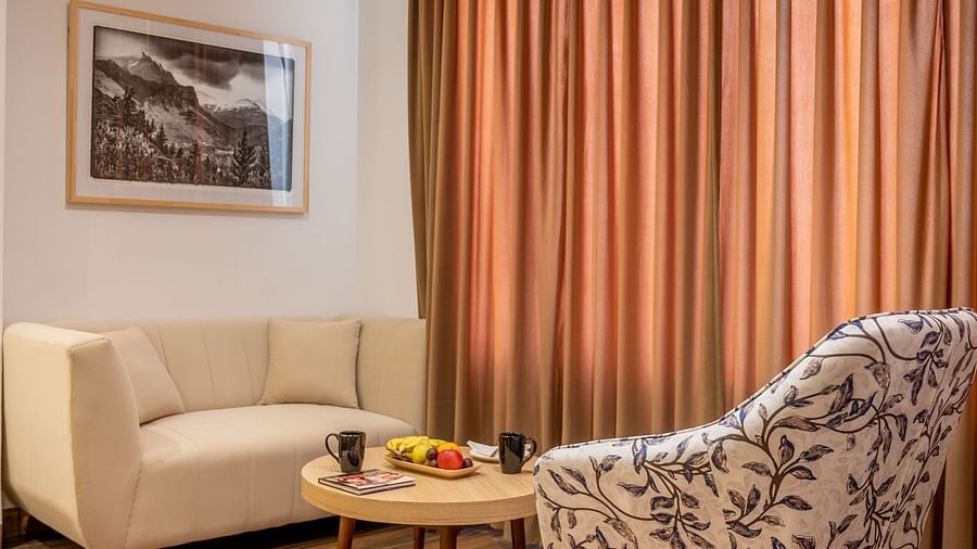 Side view of a living area of a suite at Vishranti Hotel Dehradun featuring a cushioned chair, a round table and a large window with curtains closed.