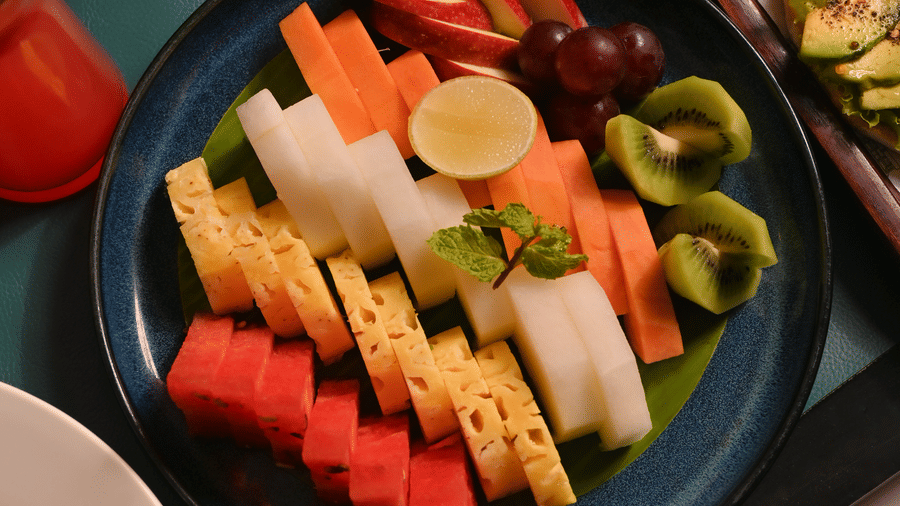 A colourful assortment of sliced fresh fruits including watermelon, papaya, pineapple, kiwi, and grapes arranged on a dark plate, with avocado slices and other breakfast items nearby on a tray - Ananta Spa and Resort, Pushkar.