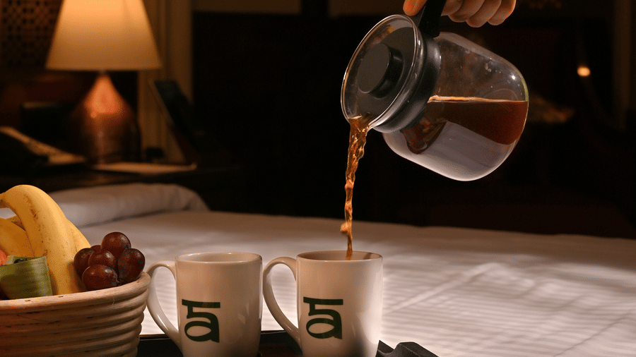 A person pouring coffee from a glass carafe into a mug on a tray with assorted breakfast items, including fruit and condiments, set on a bed in a cosy, warmly lit room with a bedside lamp in the background - Ananta Spa and Resort, Pushkar.