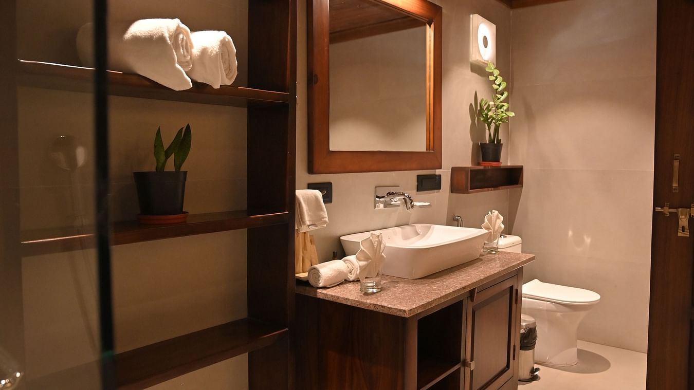 A washroom of Premium Tent featuring a wooden shelf where some folded towels are placed, a cabinet and above it  there is a sink and faucet and a mirror attached on the wall - The Baagh Ananta Elite, Ranthambore