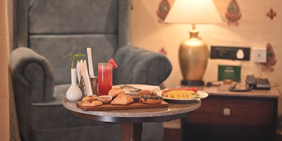 A Premium Tent featuring a sofa chair, a centre table which has a tray with croissant, cookies, doughnuts, a glass of juice and near to it there is a bedside table  with lamp and a telephone - The Baagh Ananta Elite, Ranthambore