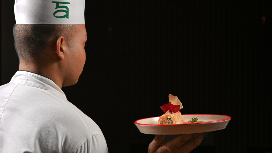 A chef wearing a tall white hat and white uniform is seen from behind, holding a plated gourmet dish with careful presentation, against a dark background - Ananta Spa and Resort, Pushkar.