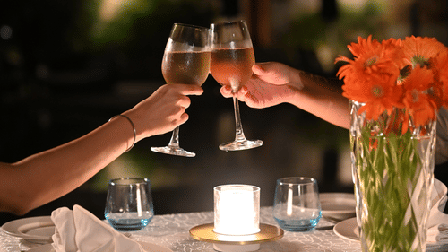Close-up of a romantic candlelight dinner setup with two people clinking wine glasses, a bouquet of orange flowers, and gourmet appetisers on a white tablecloth - Ananta Spa and Resort, Pushkar.