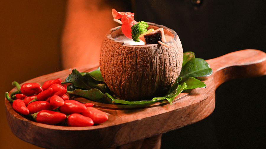 A person dressed in a black chef's uniform is holding a wooden serving board with a coconut bowl filled with food, garnished with herbs and vegetables, and accompanied by a cluster of red chili peppers - Ananta Spa and Resort, Pushkar.